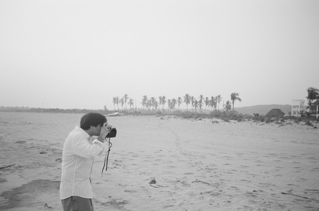 Me, standing on a beach with trees in the background, taking a photo to my right.